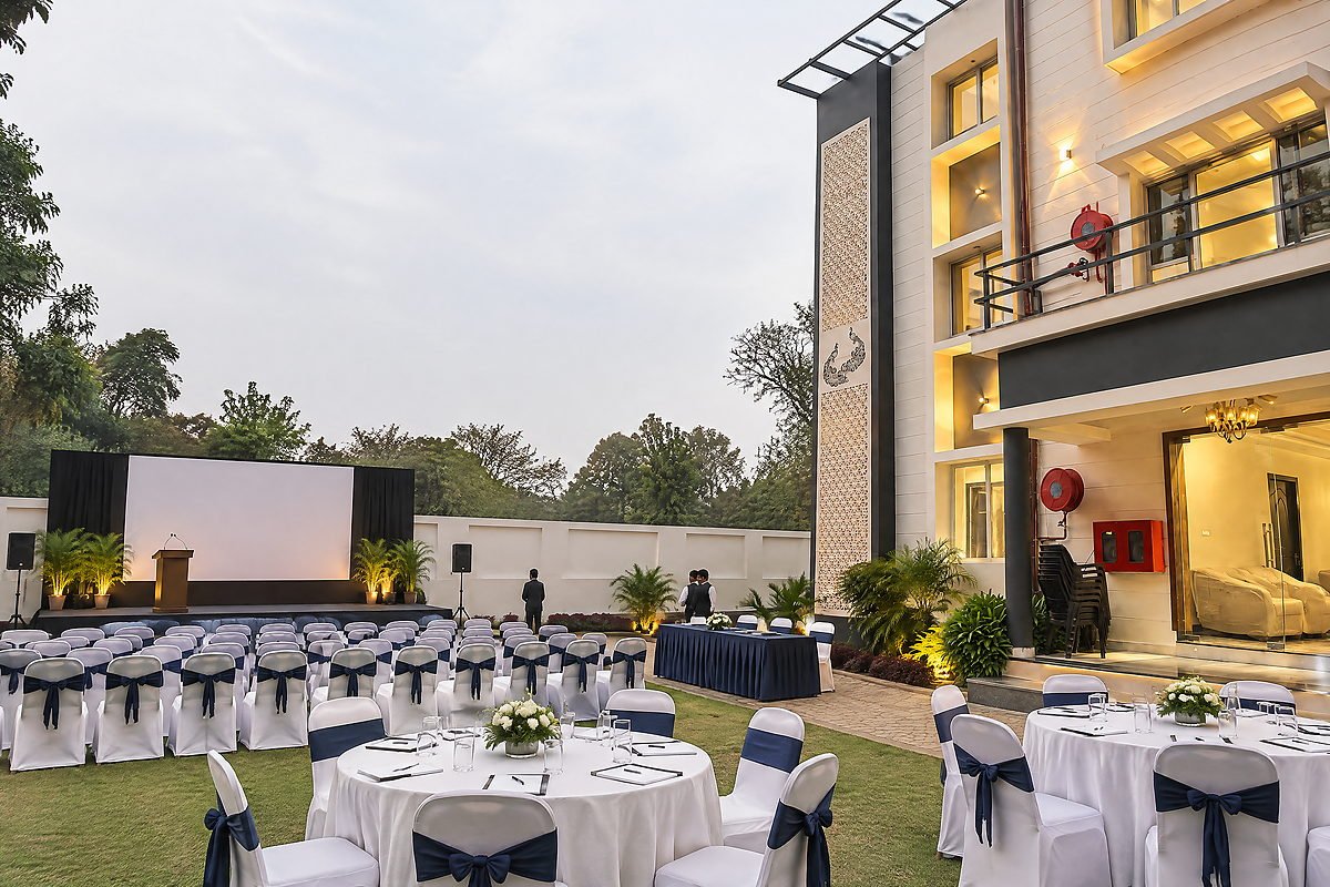 A corporate function set up at The White House Pilibhit in daylight, with rows of seating facing a podium and screen, and banquet tables behind